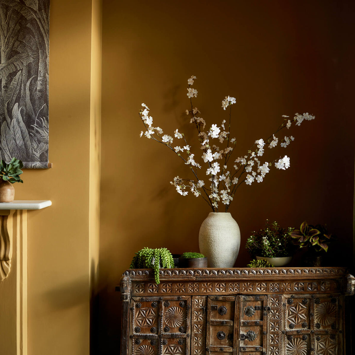 A rustic wooden cabinet holds a beige vase with white blossoms and several potted plants, set against a warm brown wall with soft natural light and botanical wall art nearby.