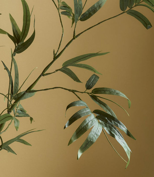 A close-up of the 110 cm Faux Bamboo Stem, Green, shows thin green leaves and stems against a muted beige background, with soft lighting that highlights the texture and evokes subtle Japanese tranquillity.