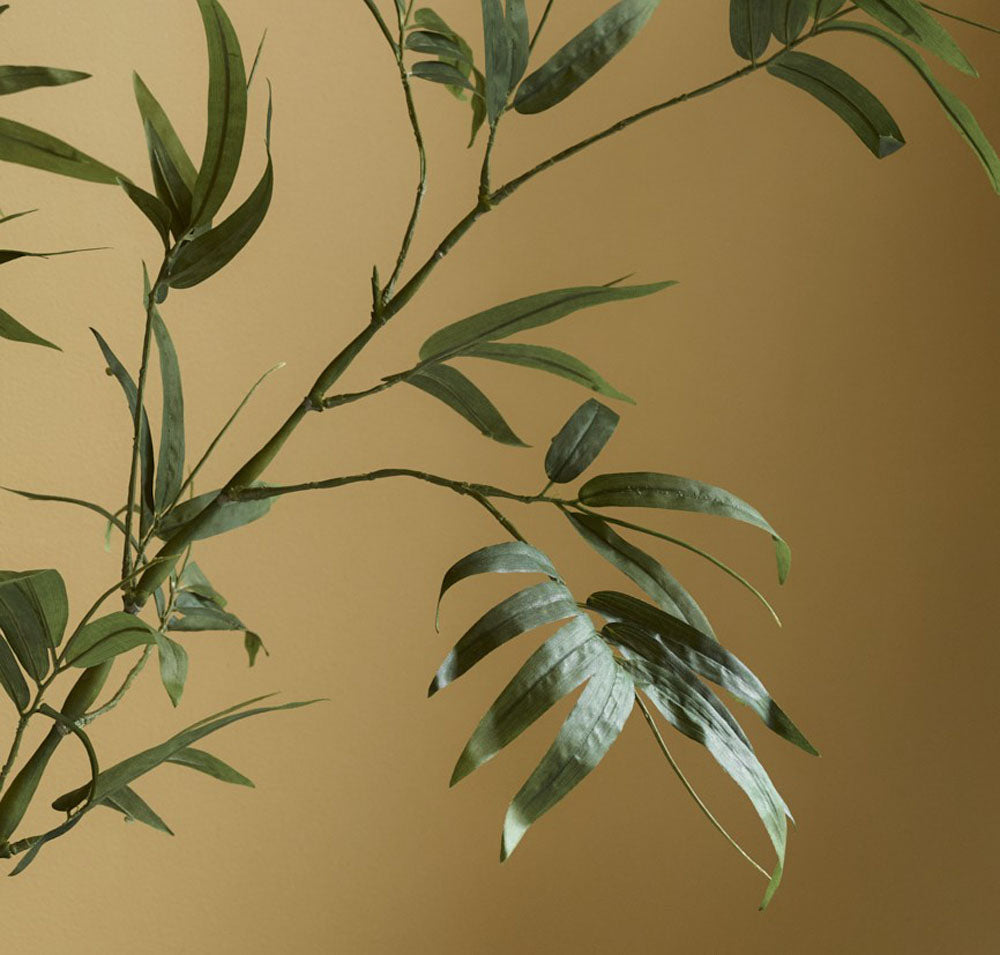 A close-up of the 110 cm Faux Bamboo Stem, Green, shows thin green leaves and stems against a muted beige background, with soft lighting that highlights the texture and evokes subtle Japanese tranquillity.