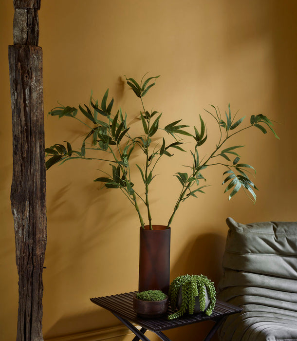 A Faux Bamboo Stem, Green - 110 cm stands in a brown vase on a small black table with a potted succulent, beside a green cushioned chair in a yellow-walled room with rustic wood, evoking Japanese tranquillity.