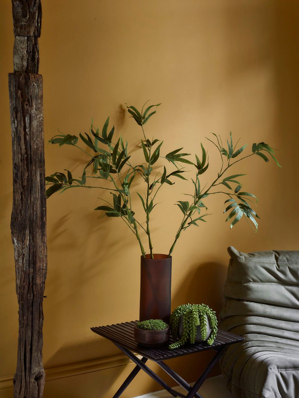 A Faux Bamboo Stem, Green - 110 cm stands in a brown vase on a small black table with a potted succulent, beside a green cushioned chair in a yellow-walled room with rustic wood, evoking Japanese tranquillity.