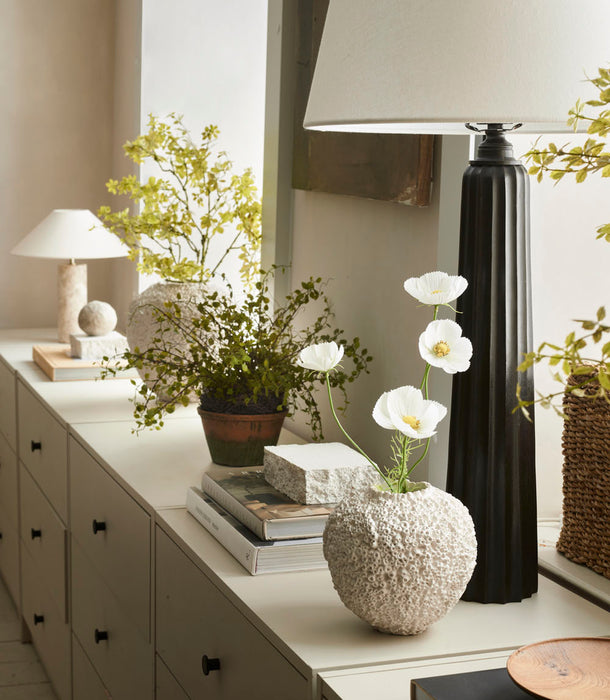 A modern sideboard displays a black lamp, the Beehive Vase by 101 Copenhagen (Birch, 16 x 16 cm) with white flowers, potted greenery, stacked books, and a woven basket. Further along are a smaller lamp and more plants, highlighting Scandinavian style.