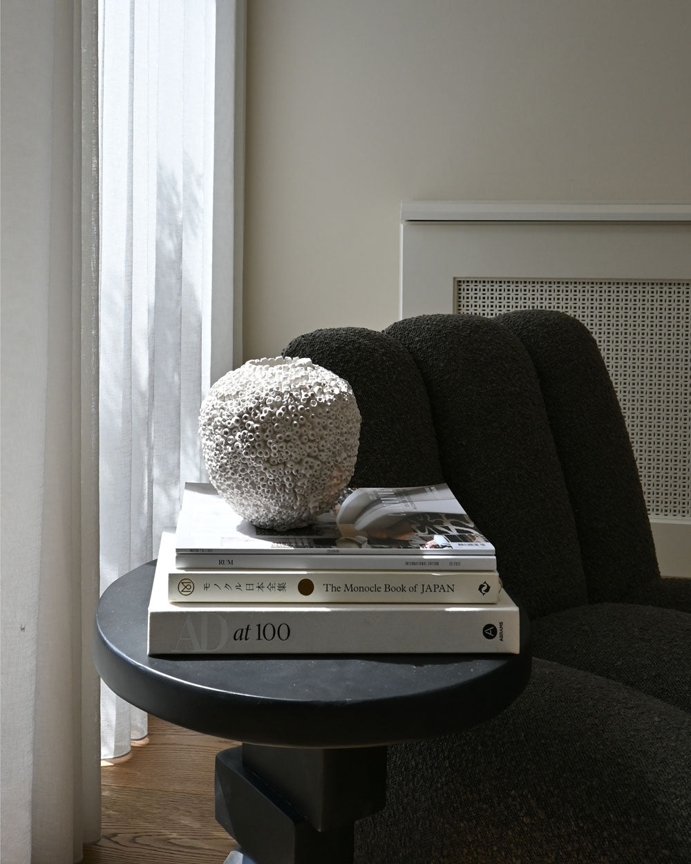 The Beehive Vase by 101 Copenhagen in Birch (16 x 16 cm) rests on a stack of books on a round black side table beside a dark chair, bathed in soft daylight through sheer white curtains, evoking understated Scandinavian style.