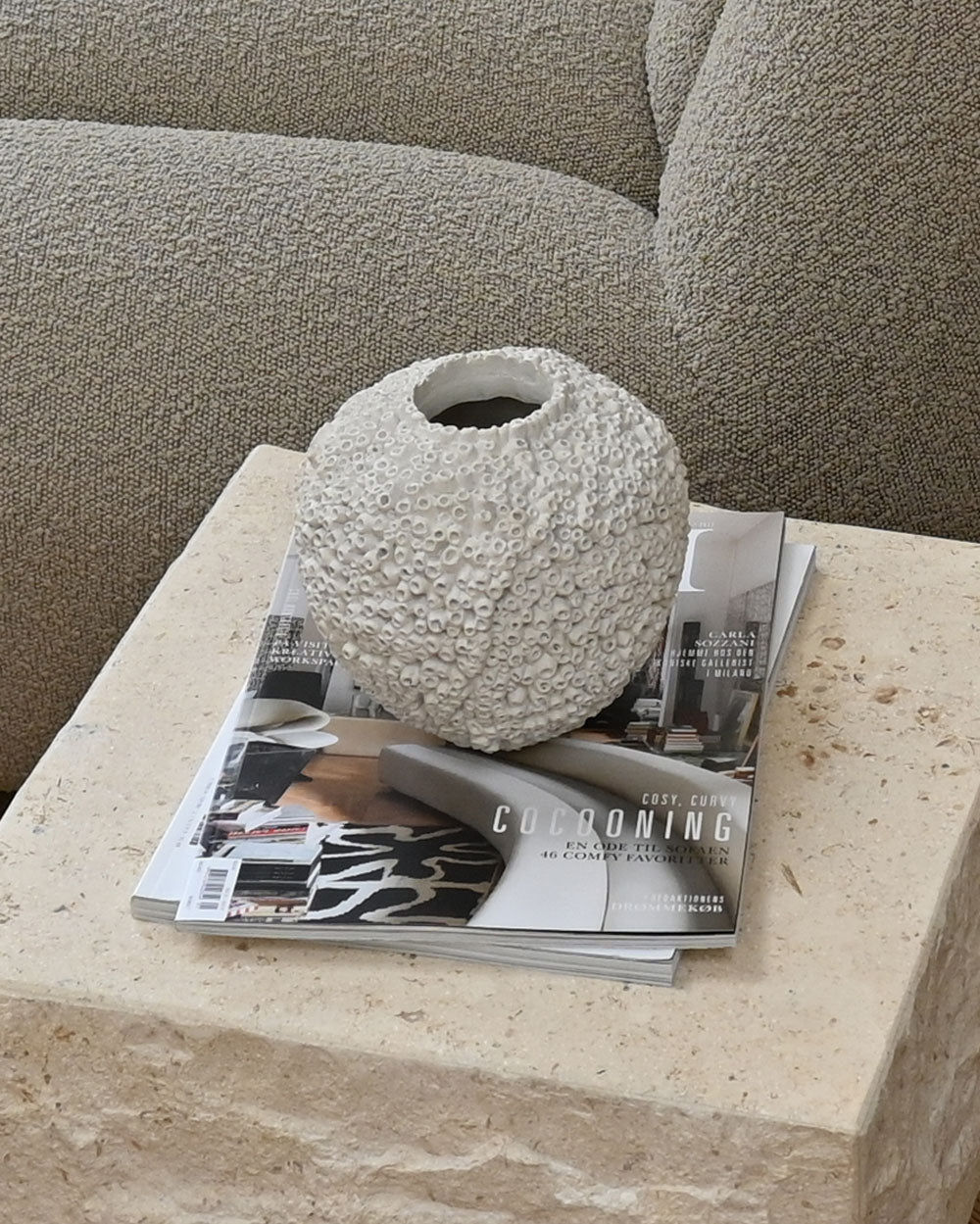 The Beehive Vase by 101 Copenhagen in birch (16 x 16 cm) sits atop magazines on a beige stone surface, echoing Scandinavian style, with a light gray upholstered sofa in the background.