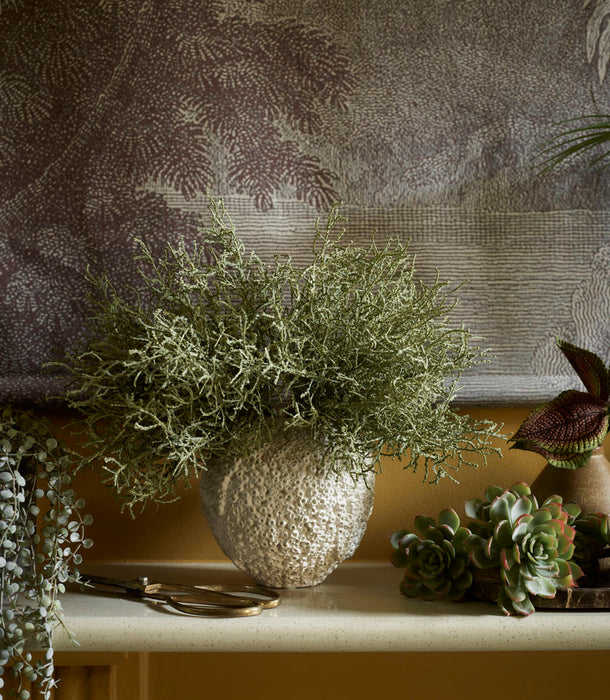 The Beehive Vase by 101 Copenhagen, Birch - 16 x 16 cm, holds wispy green foliage on a mantle, complemented by small succulents, trailing plants, and a large nature-themed tapestry in the background.