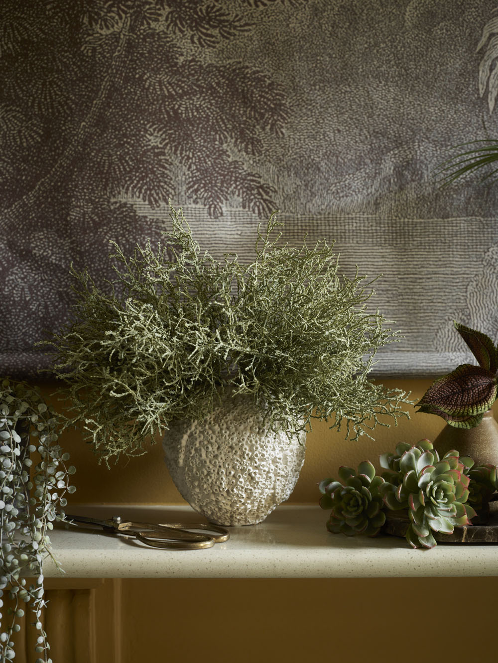 The Beehive Vase by 101 Copenhagen, Birch - 16 x 16 cm, holds wispy green foliage on a mantle, complemented by small succulents, trailing plants, and a large nature-themed tapestry in the background.