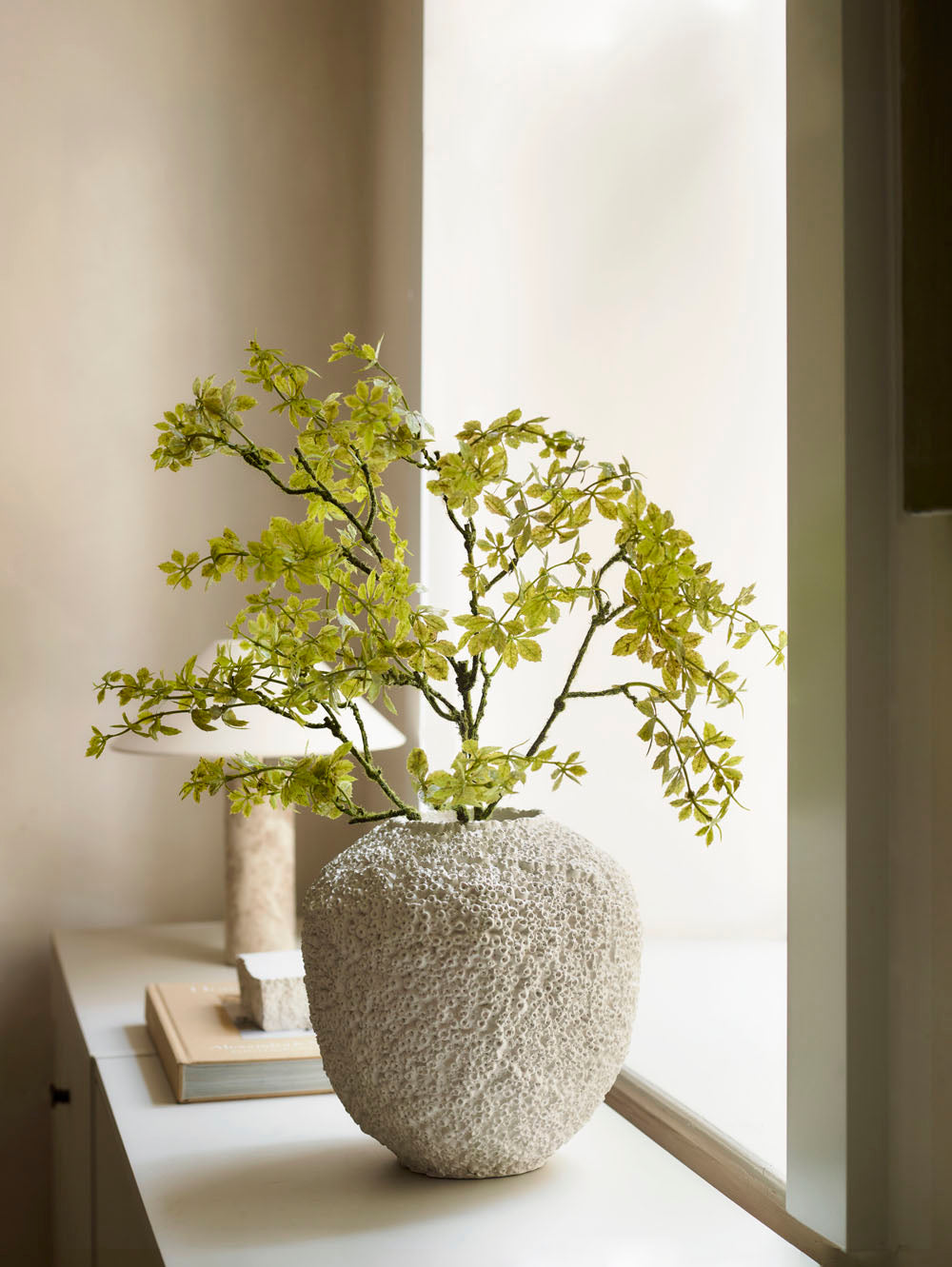 The Beehive Vase by 101 Copenhagen in Birch (25 x 25 cm) sits on a white windowsill with green branches, next to stacked books and a small decor piece, evoking serene Scandinavian charm in soft natural light.