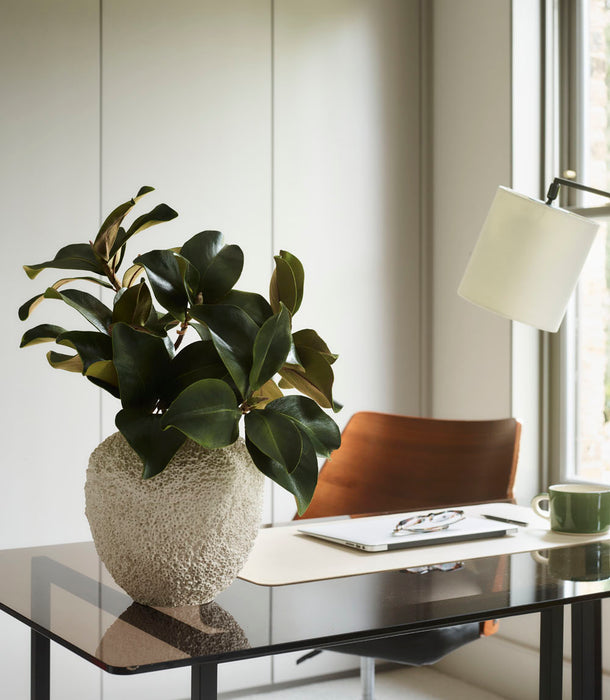 A Beehive Vase by 101 Copenhagen, Birch - 25 x 25 cm, sits on a glass desk in a modern office with Scandinavian decor. Behind the desk are a wooden chair, laptop, green mug, and white desk lamp near a window.