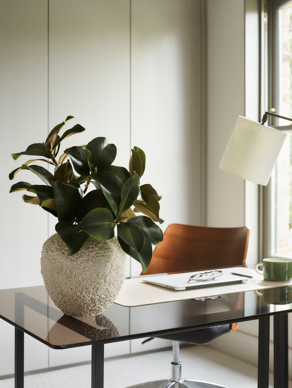 A Beehive Vase by 101 Copenhagen, Birch - 25 x 25 cm, sits on a glass desk in a modern office with Scandinavian decor. Behind the desk are a wooden chair, laptop, green mug, and white desk lamp near a window.