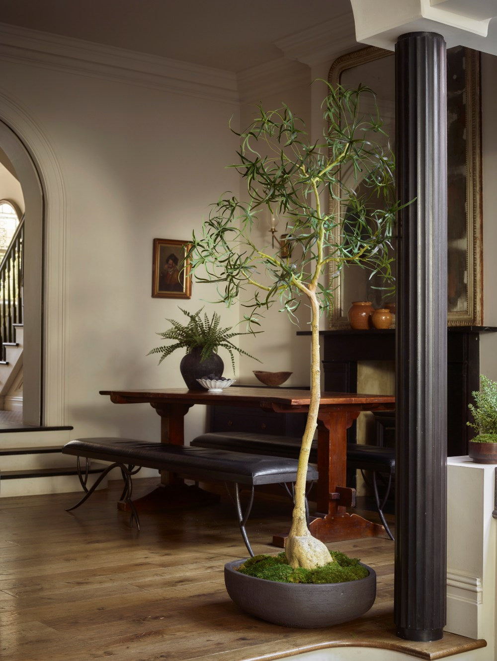 Tall faux green bottle tree displayed in a low, rounded grey planter with moss covering the base, bringing height and sculptural greenery to a traditional dining room with wooden furniture.