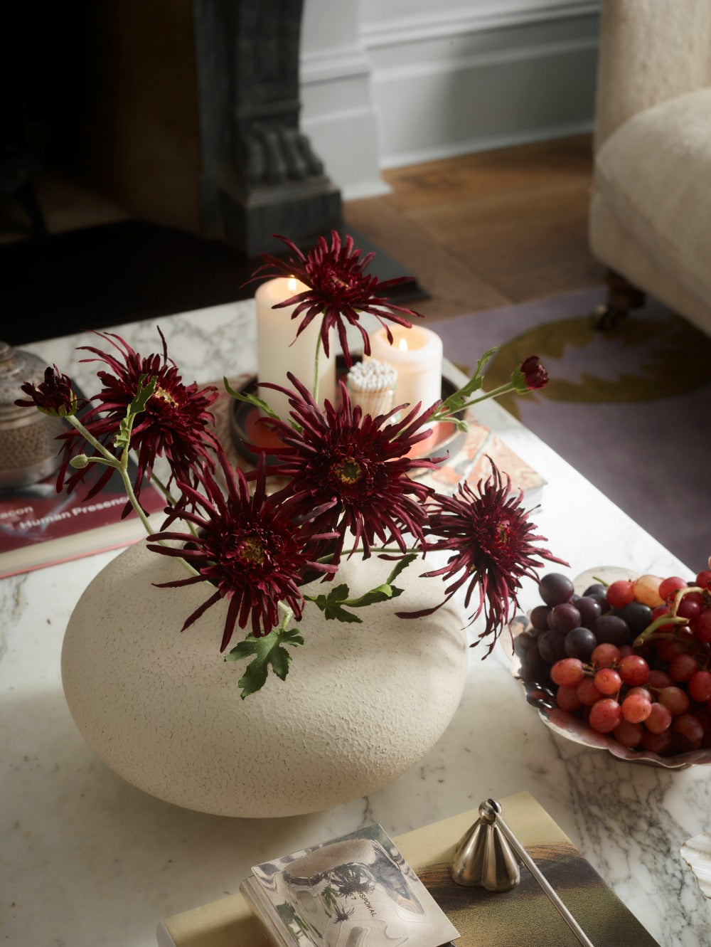Burgundy Faux Spider Chrysanthemums by Fake It Flowers, arranged in a cream ceramic vase on a marble table with decorative accessories and grapes.