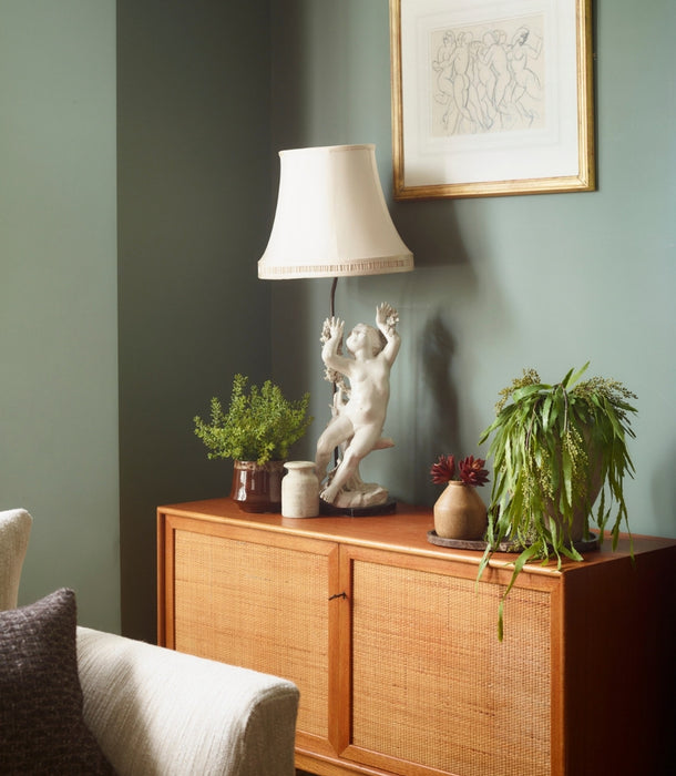 A mid-century modern wooden credenza with woven cane doors, styled in a living room setting with a table lamp, an artificial Sedum Bush potted plant, a artificial trailing berberis plant, and a framed print on a sage green wall.