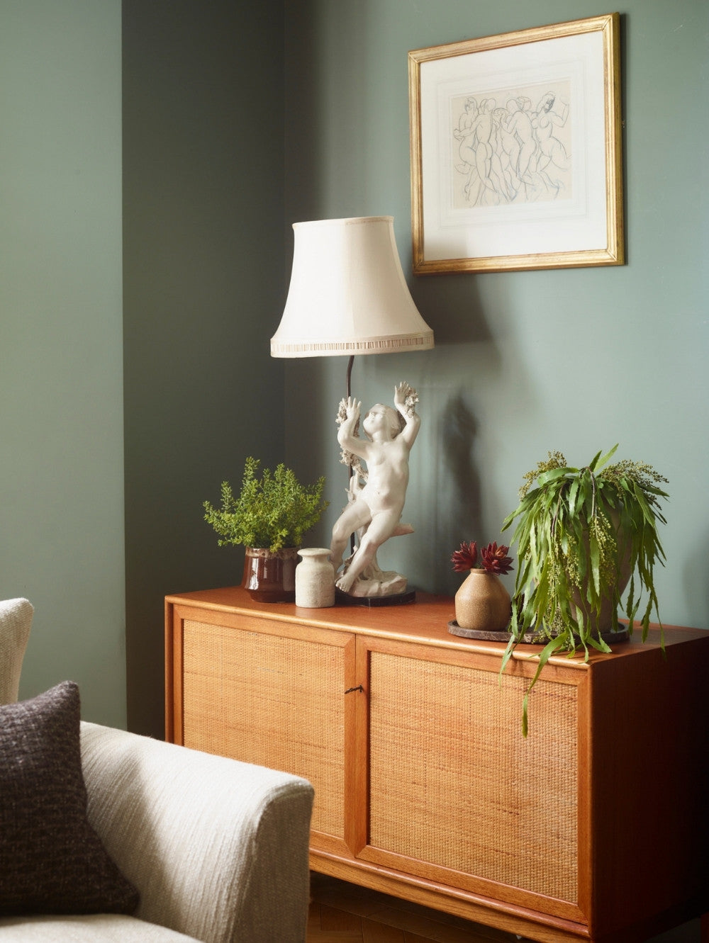 A mid-century modern wooden credenza with woven cane doors, styled in a living room setting with a table lamp, an artificial Sedum Bush potted plant, a artificial trailing berberis plant, and a framed print on a sage green wall.