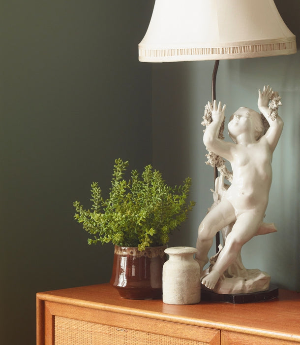 A classical-style white cherub lamp base sits on a wooden cabinet beside a Faux Sedum bush potted plant and a small vase, against a dark green wall with a lampshade visible at the top.