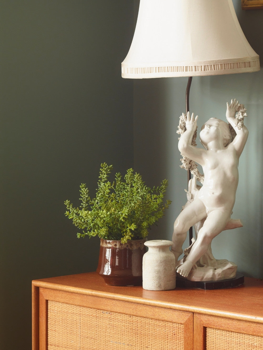 A classical-style white cherub lamp base sits on a wooden cabinet beside a Faux Sedum bush potted plant and a small vase, against a dark green wall with a lampshade visible at the top.