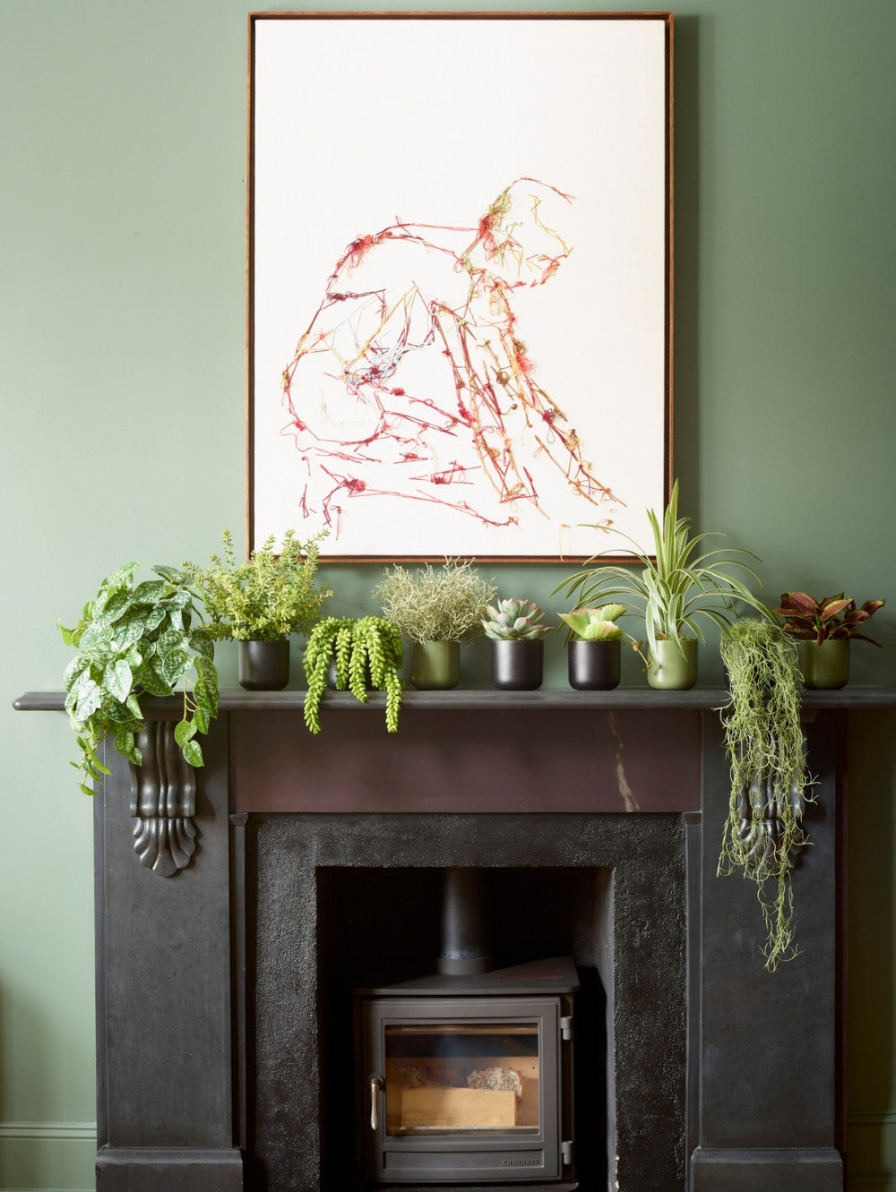 A framed abstract art print featuring red and orange line drawings on a white background, hanging on a sage green wall above a dark fireplace mantel decorated with an array of faux potted plants.