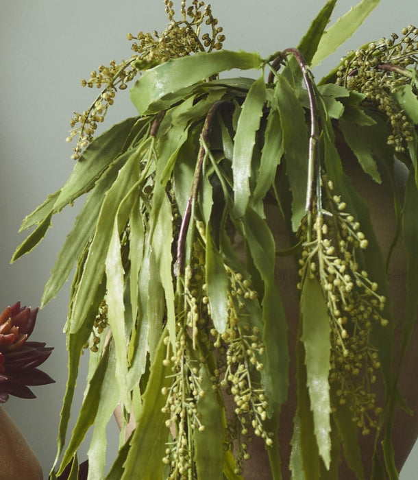 A close-up of an artificial trailing Berberis plant with long, narrow leaves and clusters of small pale green buds, hanging over a brown pot against a muted grey-green background.