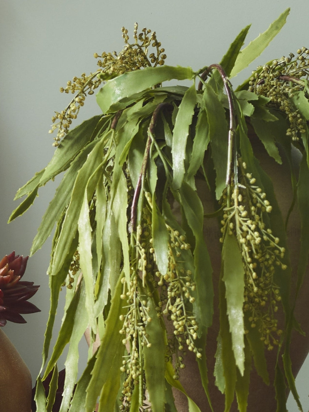 A close-up of an artificial trailing Berberis plant with long, narrow leaves and clusters of small pale green buds, hanging over a brown pot against a muted grey-green background.