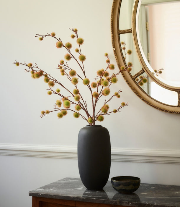 A matte black ceramic vase filled with artificial Chestnut Spray with small round yellow-green buds, displayed on a dark wood console table against a white wall with a large round gold-framed mirror.
