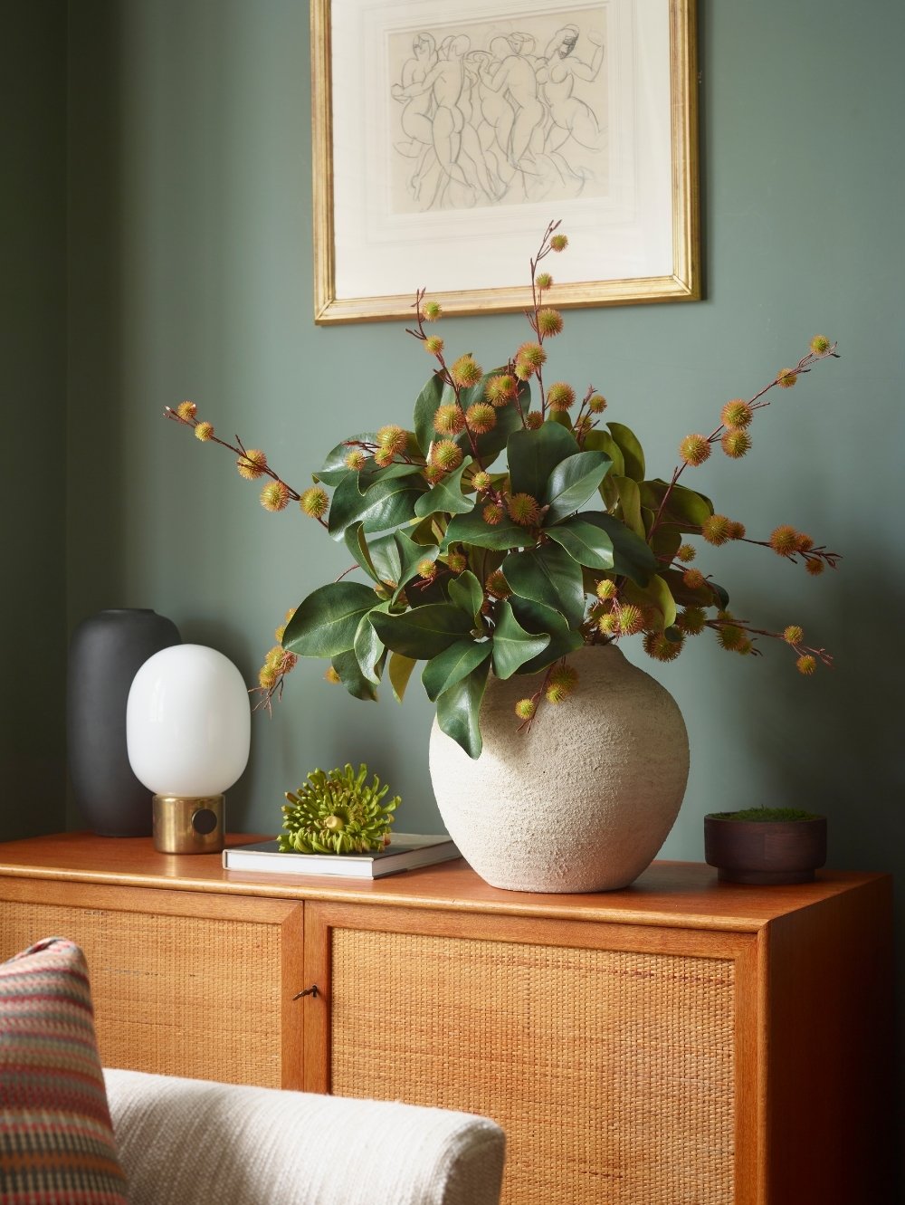 A large textured cream ceramic vase filled with green leaves and an artificial Chestnut Spray with small yellow chestnut casings, displayed on a wooden sideboard in a styled room setting.