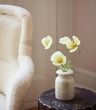A small textured ceramic vase holding four buttermilk faux cosmos flowers with green stems, placed on a dark round side table next to a cream upholstered armchair in a softly lit room.