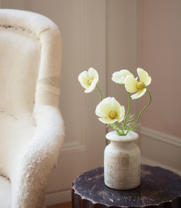 A small textured ceramic vase holding four buttermilk faux cosmos flowers with green stems, placed on a dark round side table next to a cream upholstered armchair in a softly lit room.