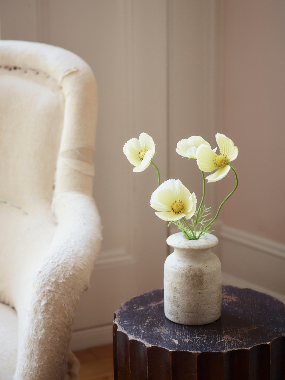 A small textured ceramic vase holding four buttermilk faux cosmos flowers with green stems, placed on a dark round side table next to a cream upholstered armchair in a softly lit room.