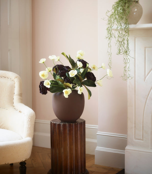 A dark brown ceramic vase filled with buttermilk faux cosmos stems and dark burgundy flowers, displayed on a dark wood pedestal in a softly lit room with pale pink walls, a cream armchair, and a fireplace mantel with artificial trailing Spanish moss greenery in a pot.