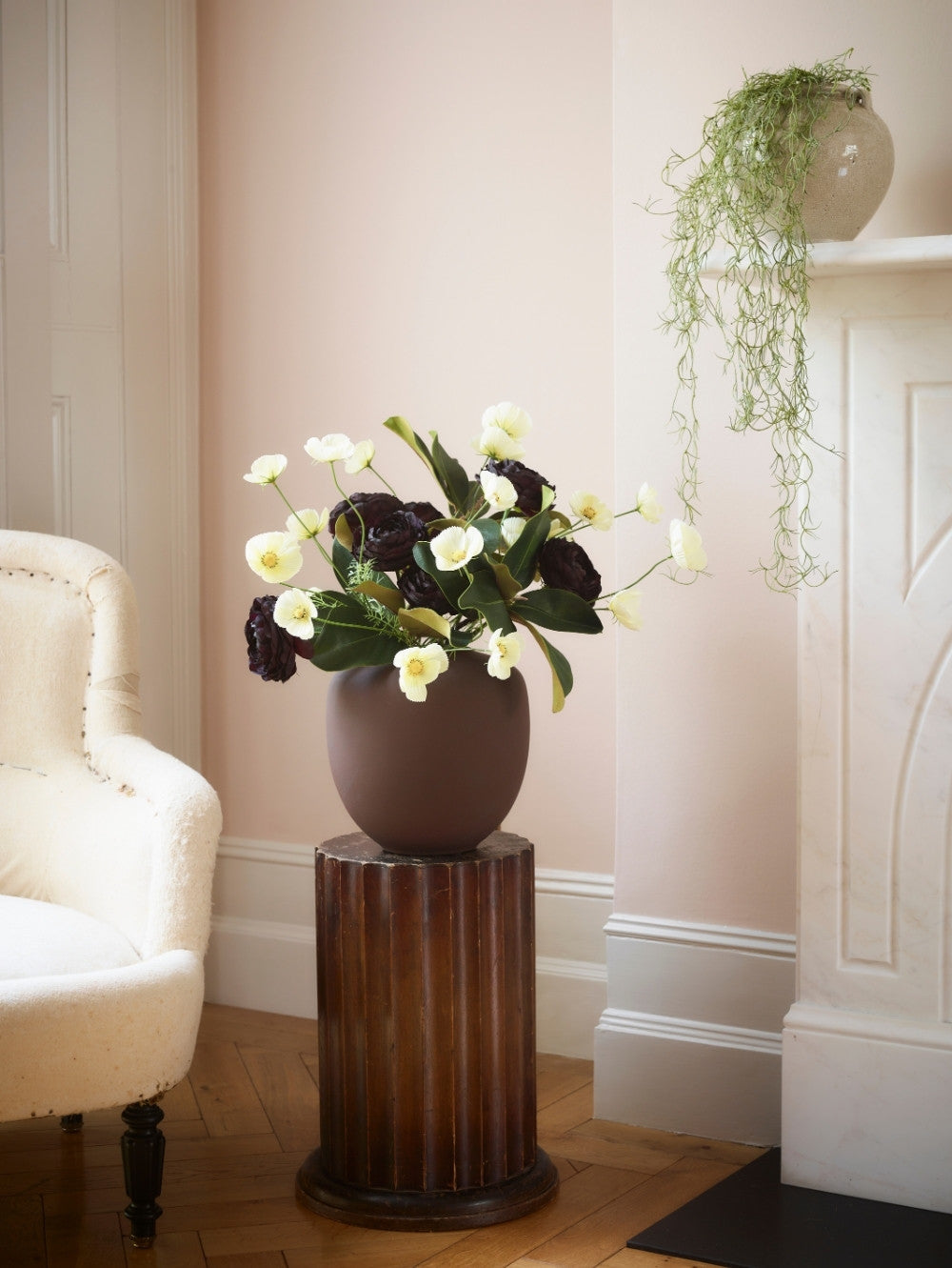 A dark brown ceramic vase filled with buttermilk faux cosmos stems and dark burgundy flowers, displayed on a dark wood pedestal in a softly lit room with pale pink walls, a cream armchair, and a fireplace mantel with artificial trailing Spanish moss greenery in a pot.