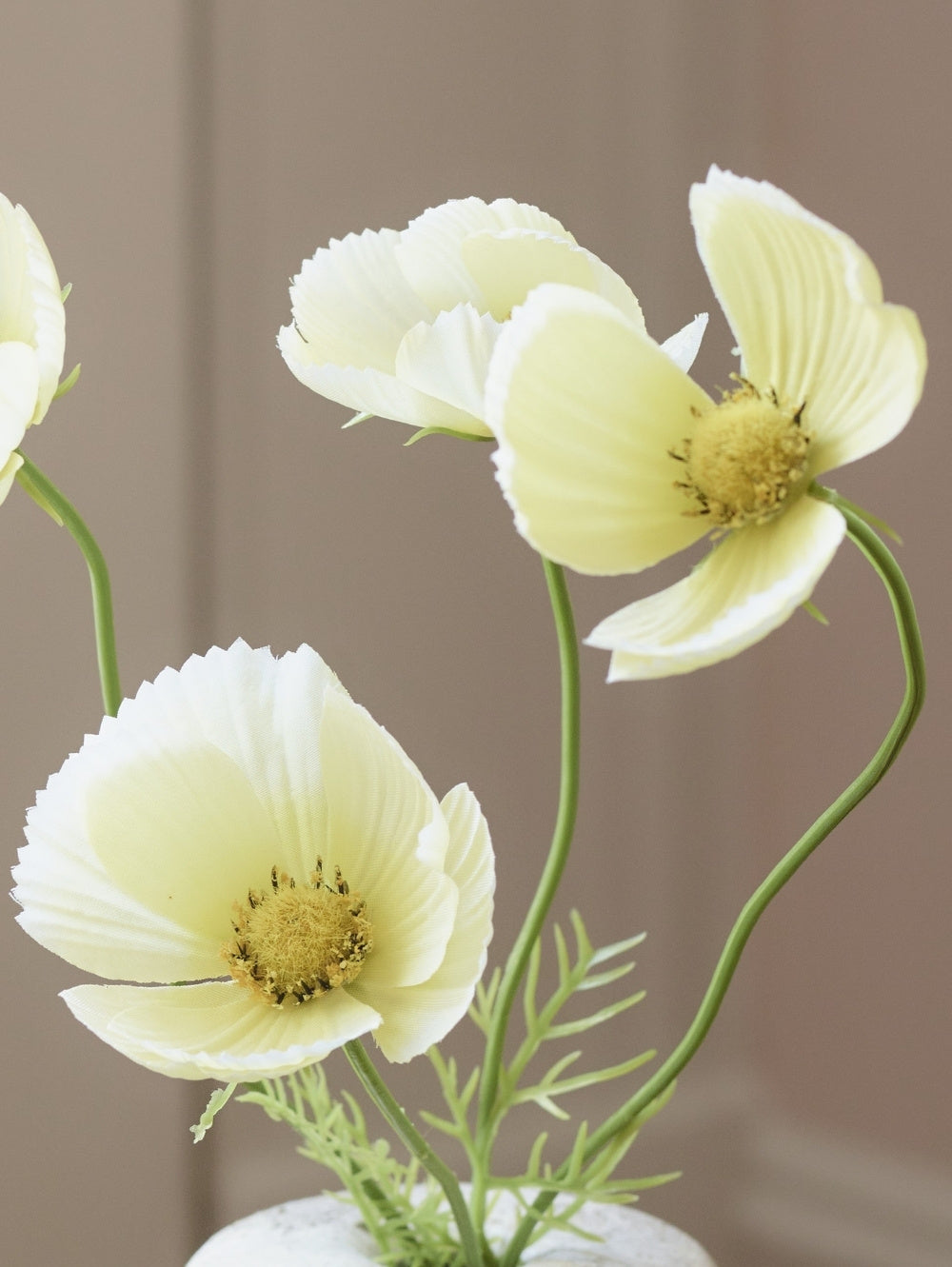 White artificial buttermilk cosmos flowers with yellow centres and green stems, arranged in a white vase against a soft beige background.