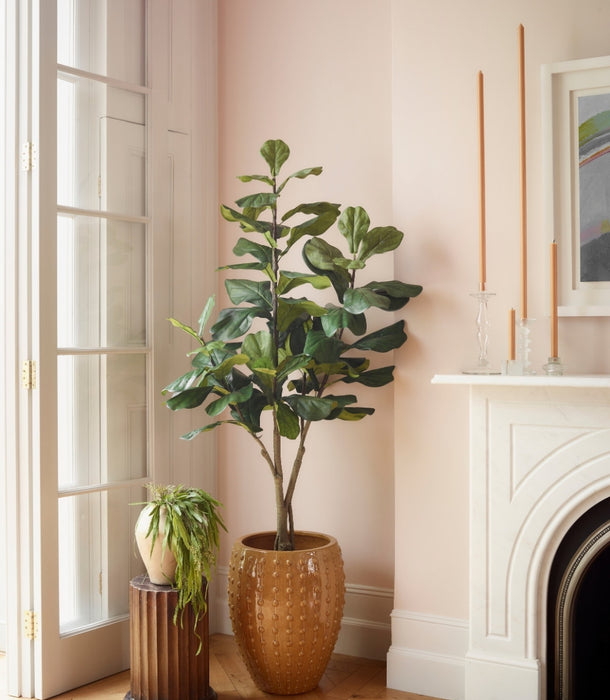 An artificial Ficus Lyrata plant 108 cm with lush green leaves stands in a textured amber-brown ceramic pot on a light wood floor, next to a white fireplace and a smaller potted plant on a dark pedestal.