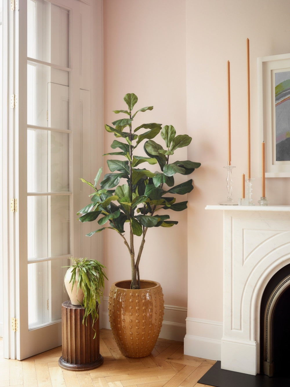 An artificial Ficus Lyrata plant 108 cm with lush green leaves stands in a textured amber-brown ceramic pot on a light wood floor, next to a white fireplace and a smaller potted plant on a dark pedestal.