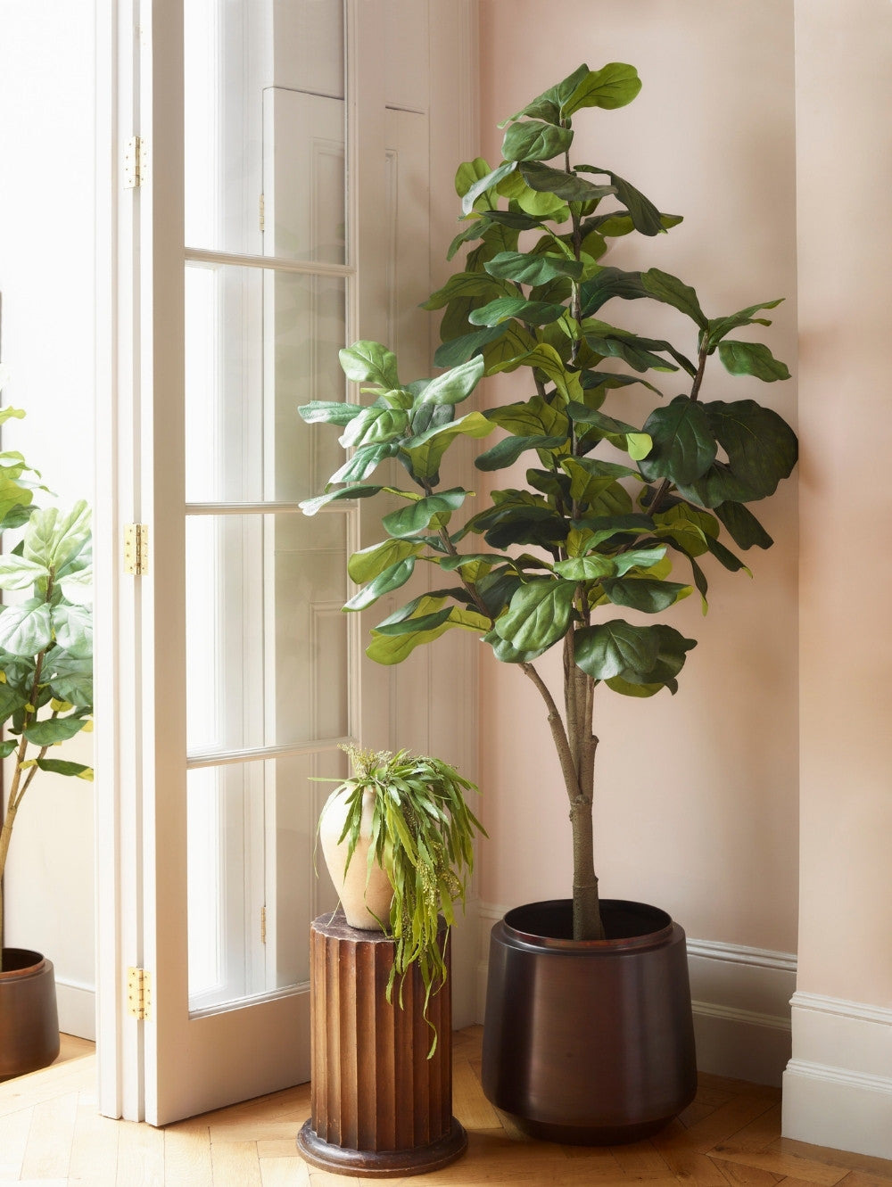 A Faux Ficus lyrata, 210 cm in a dark brown pot, standing on a wooden floor next to a cream French door, with a smaller potted trailing plant on a fluted pedestal in the background.