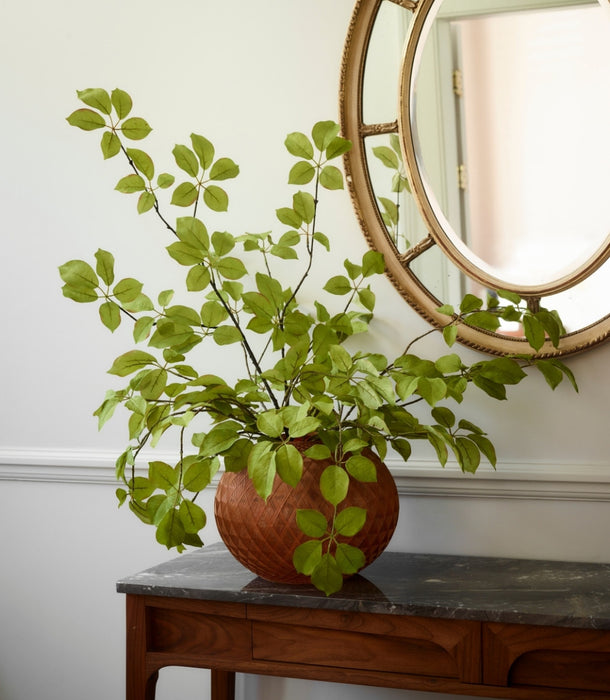 A spray of hyper-realistic faux fruit leaf branch in a textured terracotta pot, styled on a dark wood console table beneath a round gold-framed mirror against a white wall.