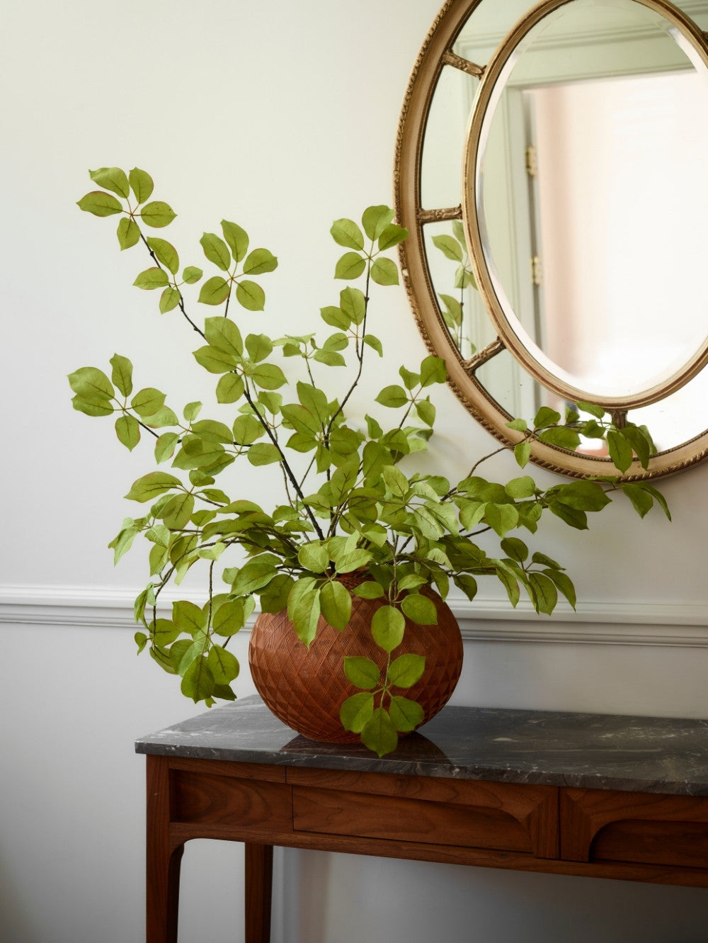 A spray of hyper-realistic faux fruit leaf branch in a textured terracotta pot, styled on a dark wood console table beneath a round gold-framed mirror against a white wall.