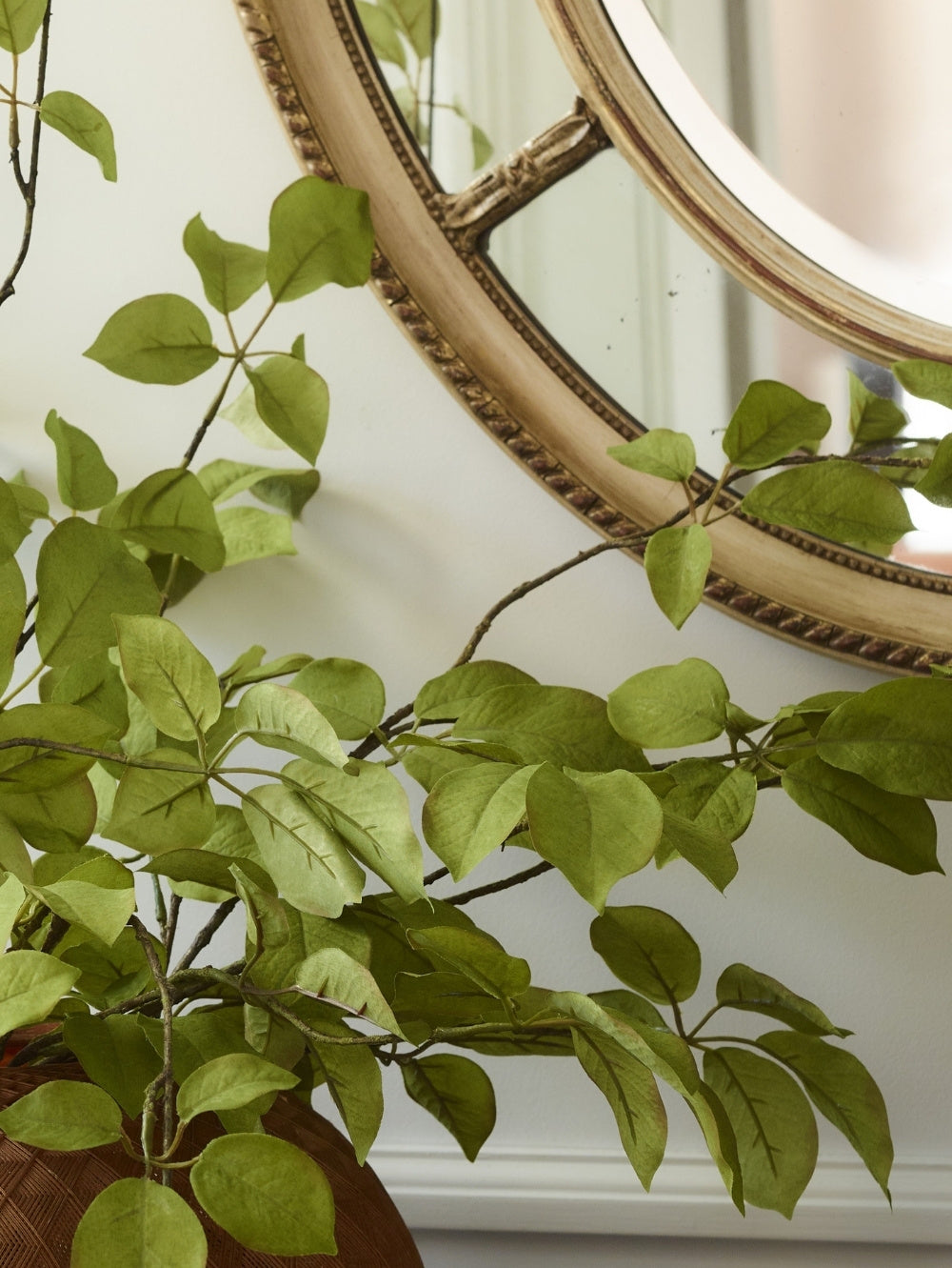 Artificial green fruit leaf branches with heart-shaped leaves draped in front of a decorative round mirror with an ornate gold frame, set against a white wall with a round panelled gold mirror