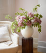 A large white ceramic vase filled with artificial pink hydrangea flowers and green leaves, displayed on a dark wood pedestal in a softly lit room next to a cream upholstered armchair.