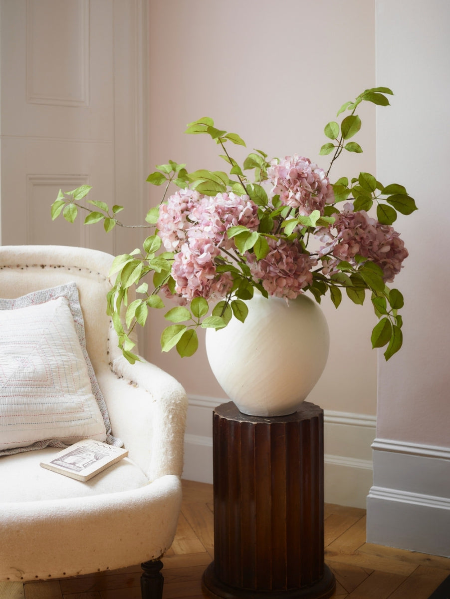 A large white ceramic vase filled with artificial pink hydrangea flowers and green leaves, displayed on a dark wood pedestal in a softly lit room next to a cream upholstered armchair.