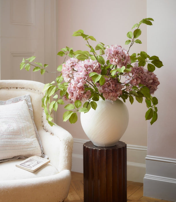 A large white ceramic vase filled with artificial pink hydrangea flowers and green leaves, displayed on a dark wood pedestal in a softly lit room next to a cream upholstered armchair.