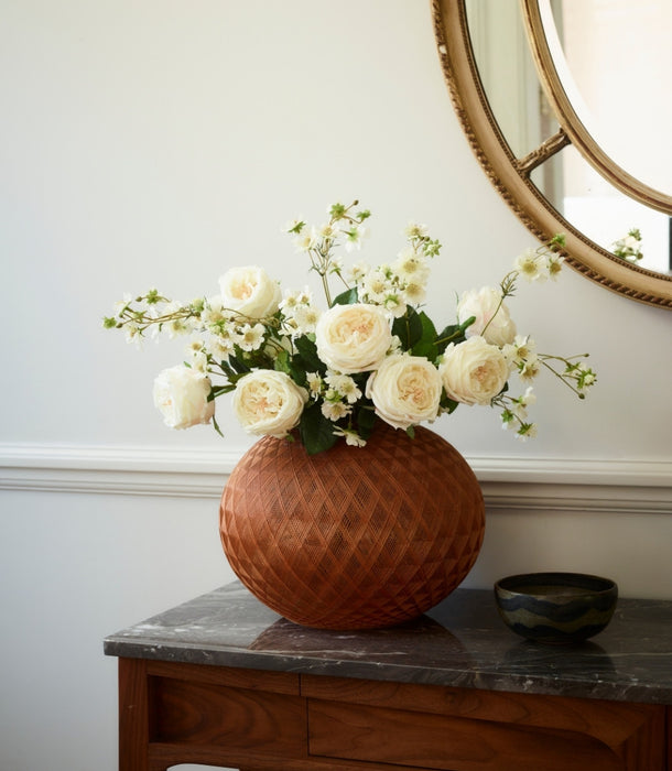 A terracotta-colored textured ceramic vase filled with creamy white artificial roses and green foliage, displayed on a dark wood console table next to a small dark bowl, with a round mirror partially visible on the wall.