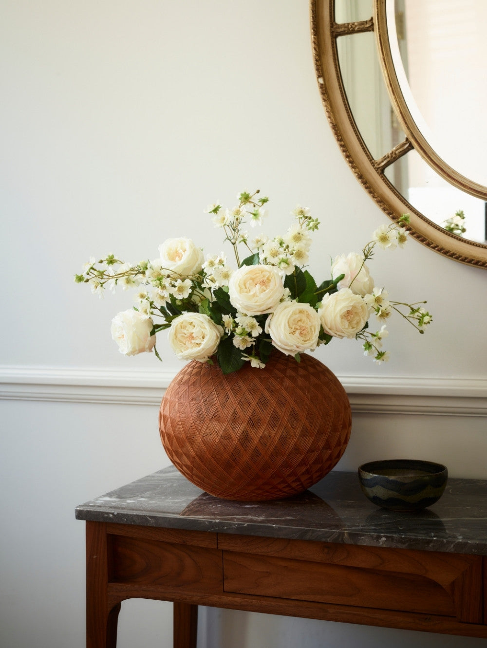 A terracotta-colored textured ceramic vase filled with creamy white artificial roses and green foliage, displayed on a dark wood console table next to a small dark bowl, with a round mirror partially visible on the wall.