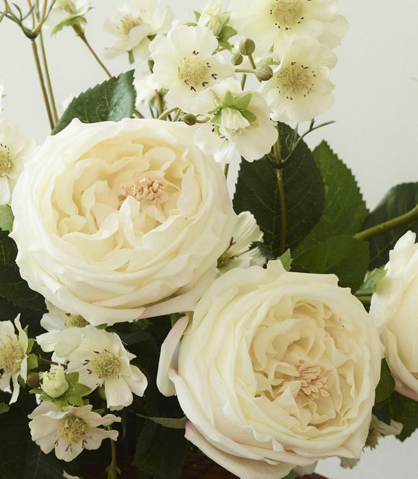 A close-up of a floral arrangement featuring large cream-colored faux roses and smaller white blossoms with green leaves, set against a plain light background.