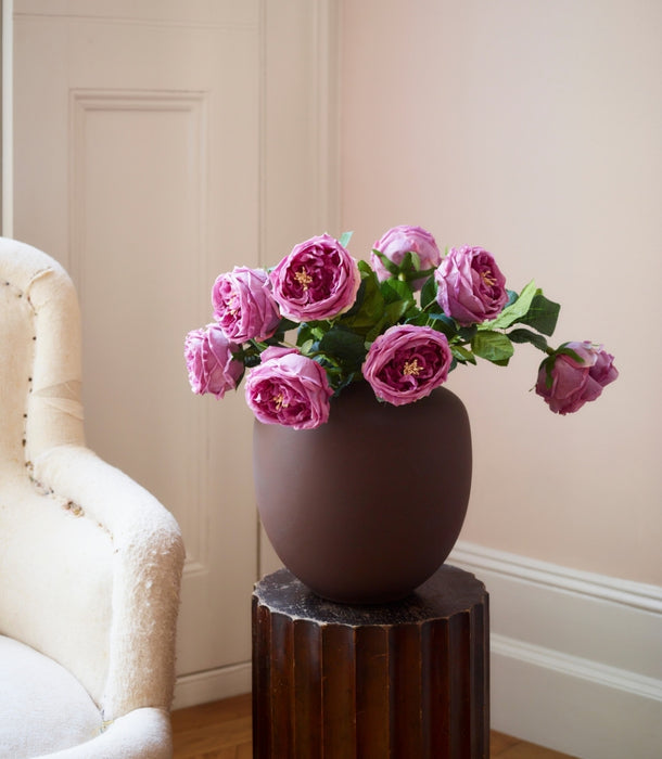 A dark brown matte ceramic vase filled with artificial pink roses, displayed on a dark wood pedestal in a softly lit room with a cream armchair and a pale pink wall.