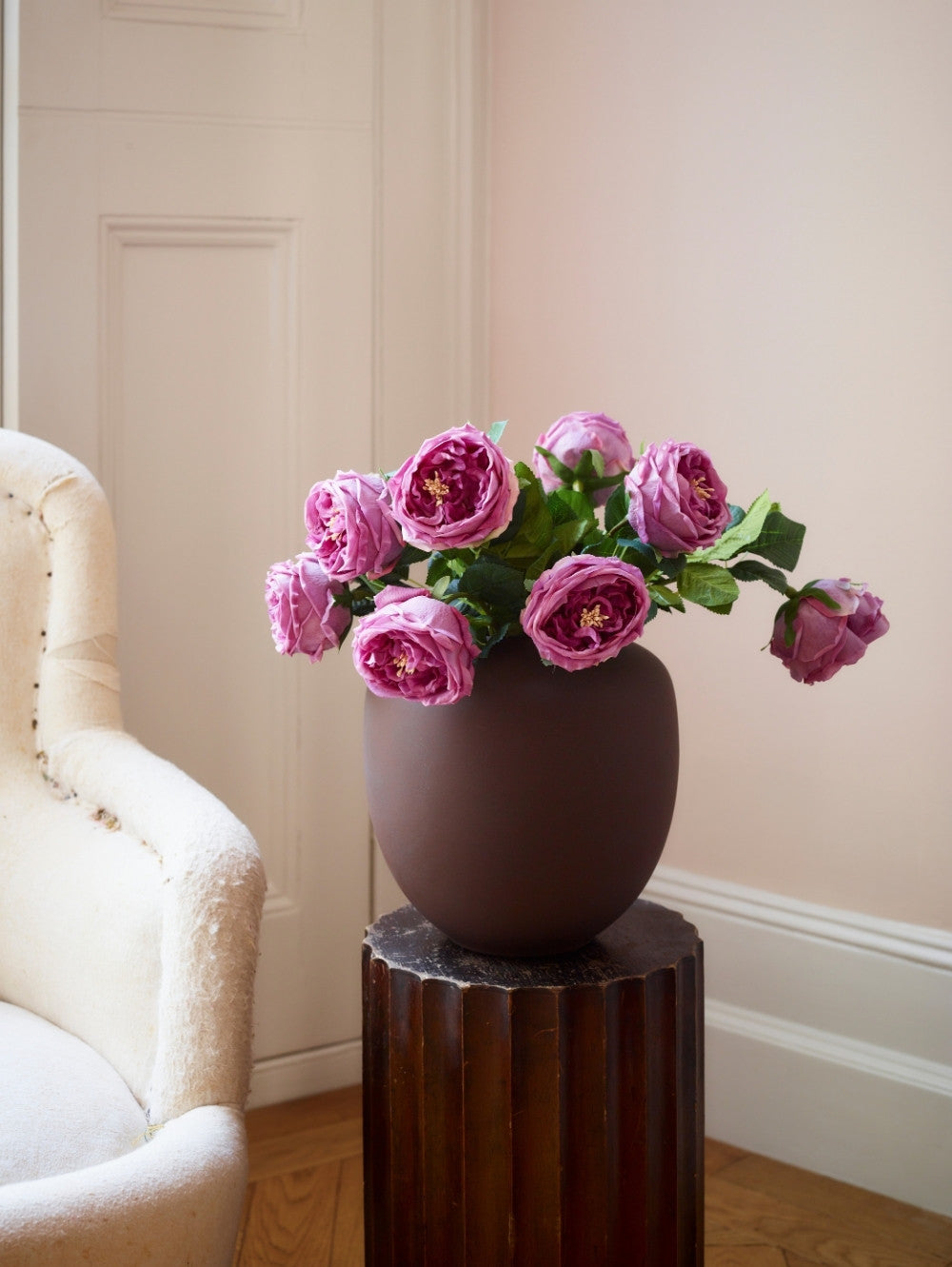 A dark brown matte ceramic vase filled with artificial pink roses, displayed on a dark wood pedestal in a softly lit room with a cream armchair and a pale pink wall.