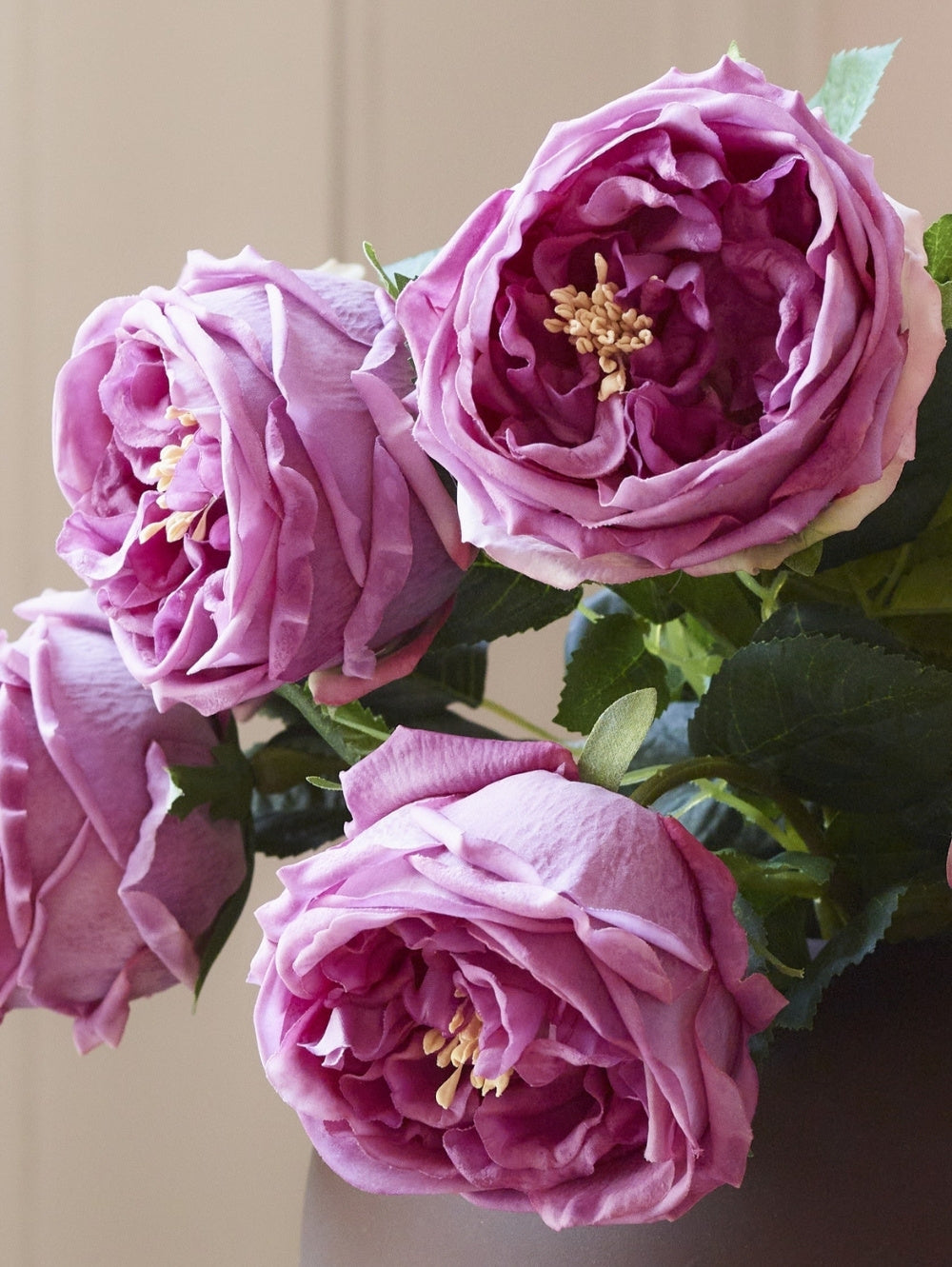 Four pinky purple artificial roses with green leaves arranged in a vase against a neutral beige background