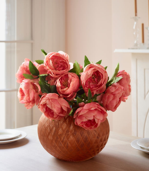 A bouquet of pinky coral faux peony flowers arranged in a textured terracotta-colored round vase, placed on a light tabletop in a softly lit interior setting.