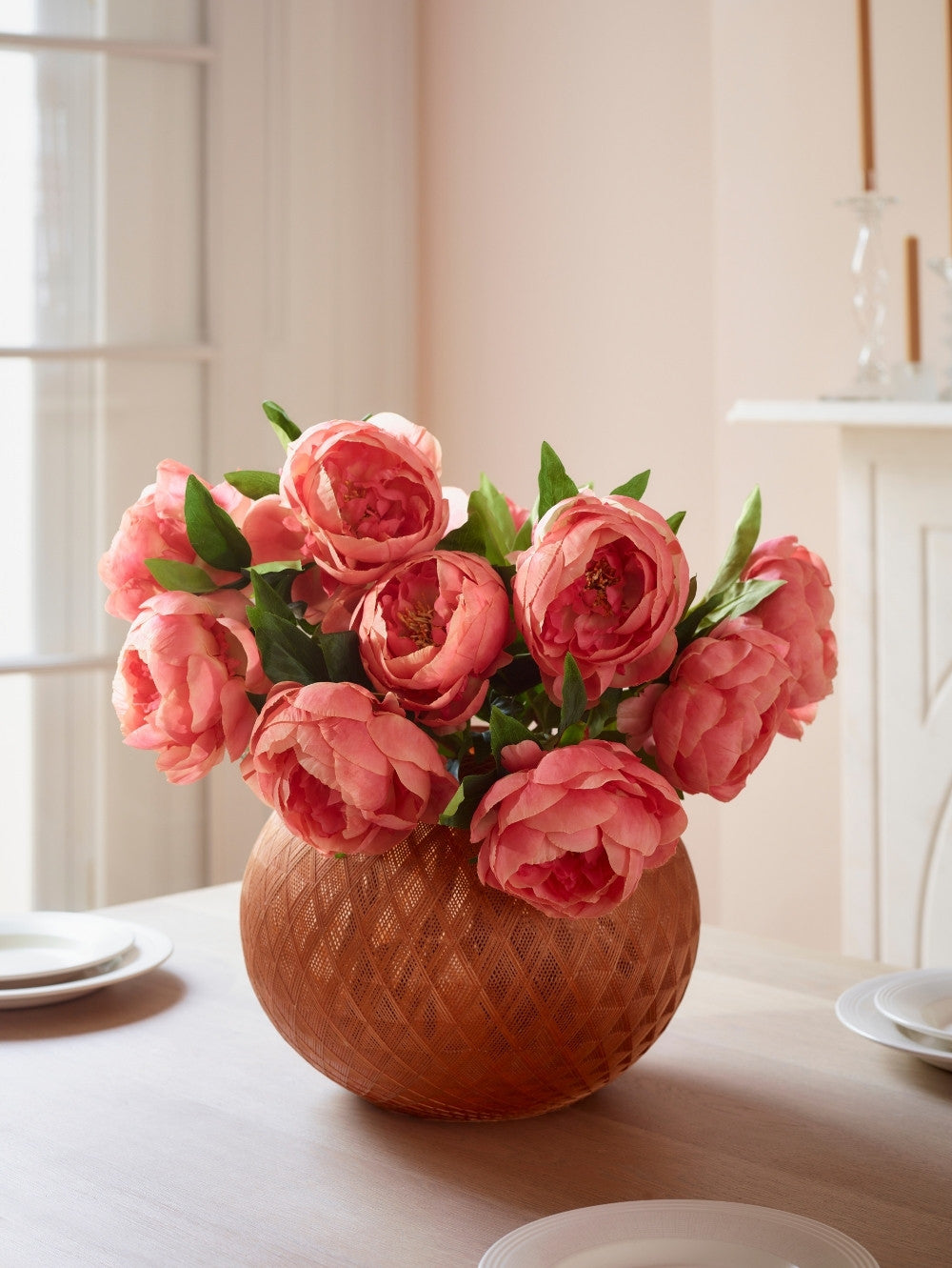 A bouquet of pinky coral faux peony flowers arranged in a textured terracotta-colored round vase, placed on a light tabletop in a softly lit interior setting.