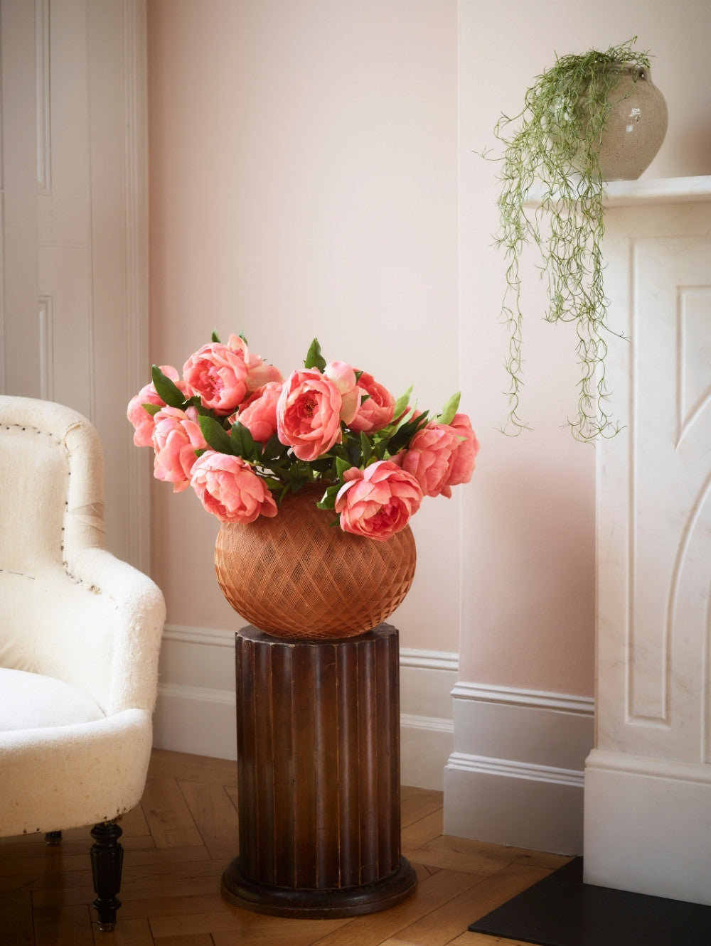 A bouquet of pink coral faux peonies in a textured terracotta pot displayed on a dark brown fluted pedestal table in a softly lit room with a cream armchair and fireplace.