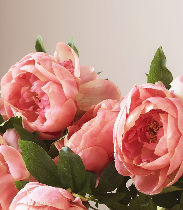A close-up of pink coral artificial peony flowers with green leaves, photographed against a soft light beige background.