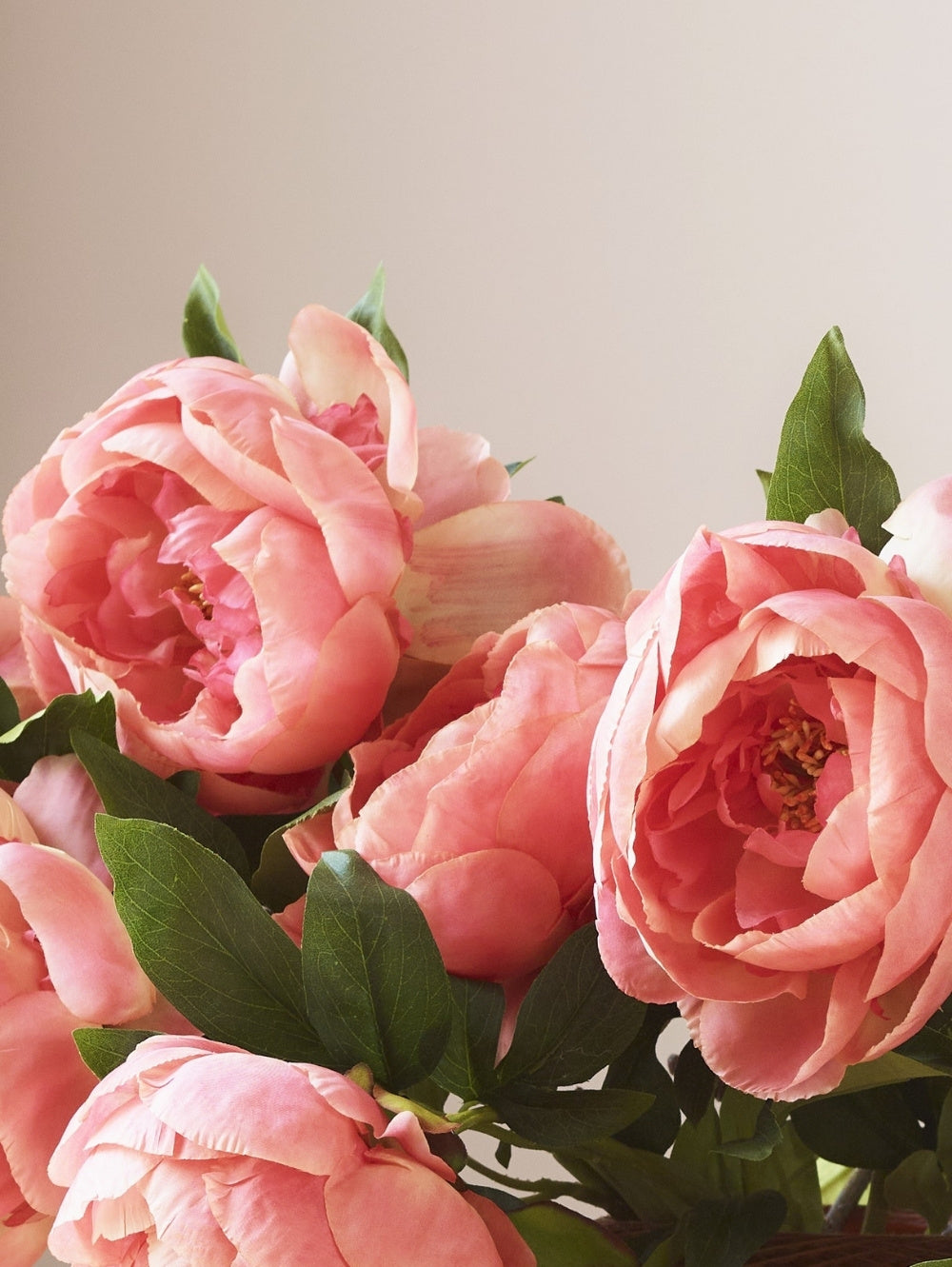 A close-up of pink coral artificial peony flowers with green leaves, photographed against a soft light beige background.
